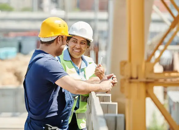 Two construction workers in hard hats smiling and talking on a building site.